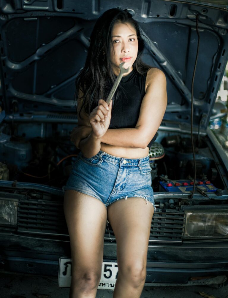 Confident woman mechanic holding a wrench standing by a car with hood open.